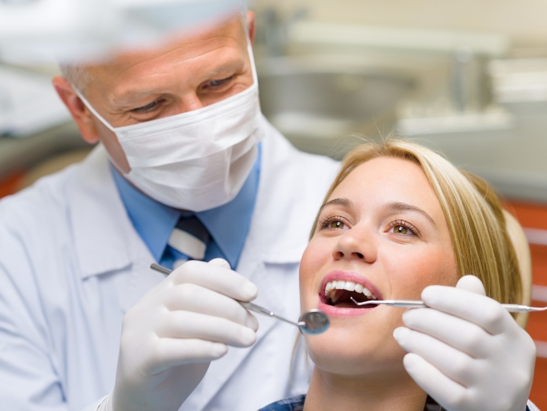 Choosing seating for long clinical days: A male dentist wearing a surgical mask and gloves examining a female patient's open mouth with a dental mirror and probe in a dental surgery.