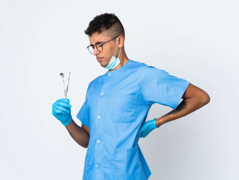 How to reduce back pain as a dentist: A male dentist in blue scrubs and gloves holding dental instruments at his side while pressing his hand to his lower back, suggesting discomfort, against a plain white background.