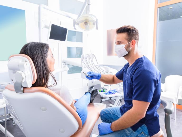 Equipment Bottlenecks in Dental Practices A dentist in navy scrubs and a face mask speaks with a patient seated in a dental chair, holding a dental instrument, with screens and equipment visible in the background.