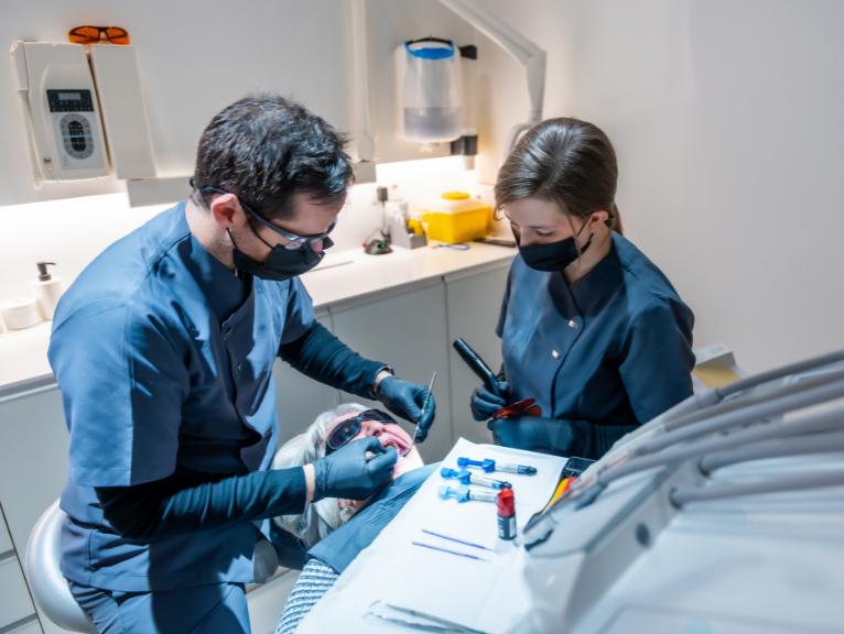How to speed up patient turnaround in dentistry A dentist and a dental nurse, both in dark scrubs and face masks, work together to treat a patient reclined in a dental chair, with instruments and syringes laid out on a tray nearby.