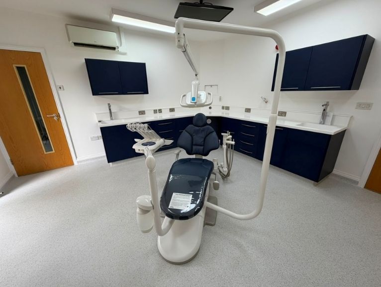 New dental practice equipment supplier: A newly fitted dental surgery room featuring a dark navy dental chair with an overhead light and arm unit, white worktops, and navy cabinetry along three walls.
