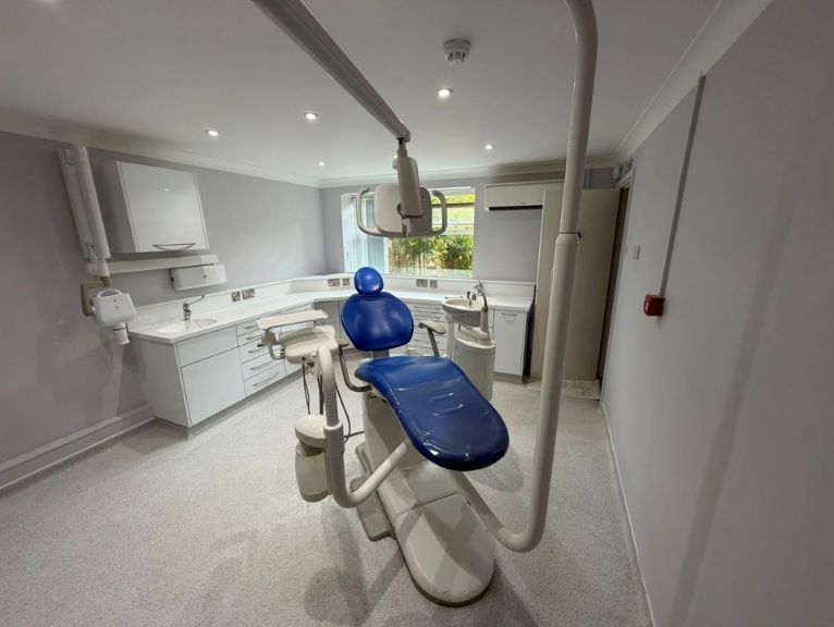 Squat dental practice fit-out: A newly fitted dental surgery room featuring a blue dental chair with an overhead light and arm unit, white worktops and cabinetry, and a window overlooking greenery.