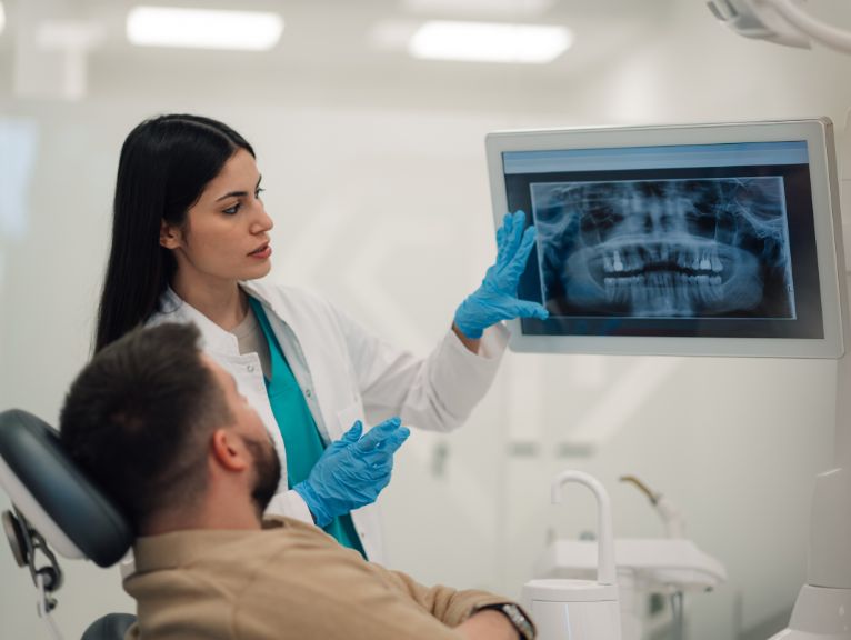 How Does Digital Imaging Help Detect Mouth Cancer Early: A dentist in a white coat and blue gloves points to a panoramic dental X-ray on a wall-mounted screen while a male patient in the dental chair looks on.