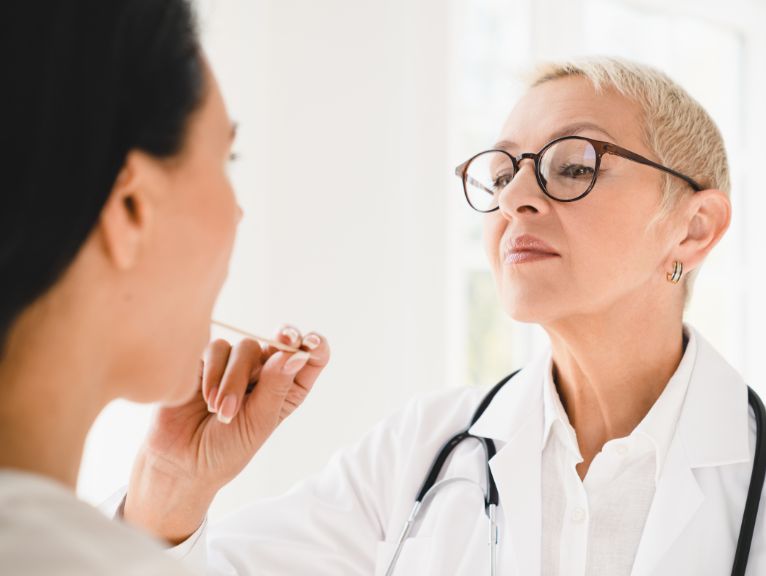 why is early detection important for mouth cancer: An older female doctor with short blonde hair, glasses and a stethoscope holds a wooden tongue depressor to examine a dark-haired woman’s mouth.